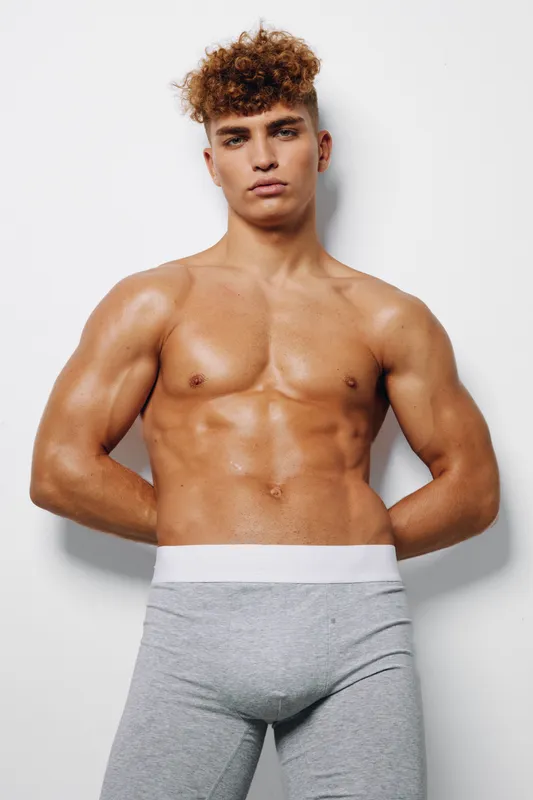 Muscular man with curly hair and athletic body posing shirtless in tight gray pants against white background, showing fitness strength and confidence in studio lighting.