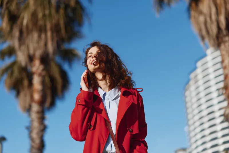 A young woman in a red coat smiles while talking on a smartphone outdoors on a sunny day with palm trees and modern buildings in the background.