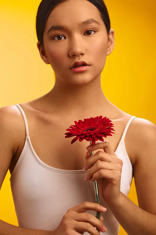 Asian woman in a simple white tank holds a red flower against a warm yellow backdrop, conveying calm poise, gentle mood, and graceful simplicity in a studio setting.