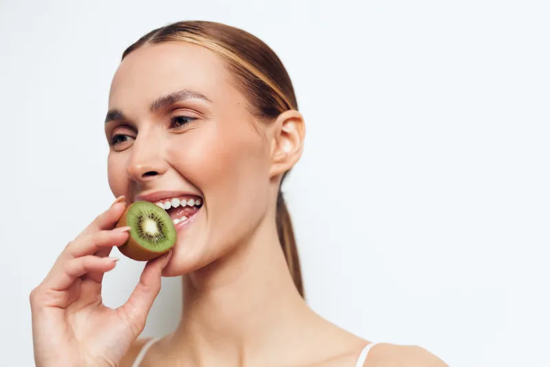 A smiling young woman with clear skin holds a slice of kiwi near her mouth on a white background. Concept for healthy lifestyle, natural beauty, and fresh fruit.