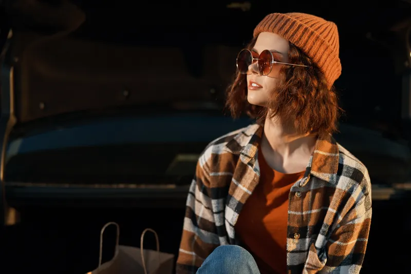 woman portrait with sunglasses and beanie by car trunk during golden hour, shopping bags and sunlit plaid jacket, candid lifestyle vibe with authenticity, mindful living and emotional storytelling