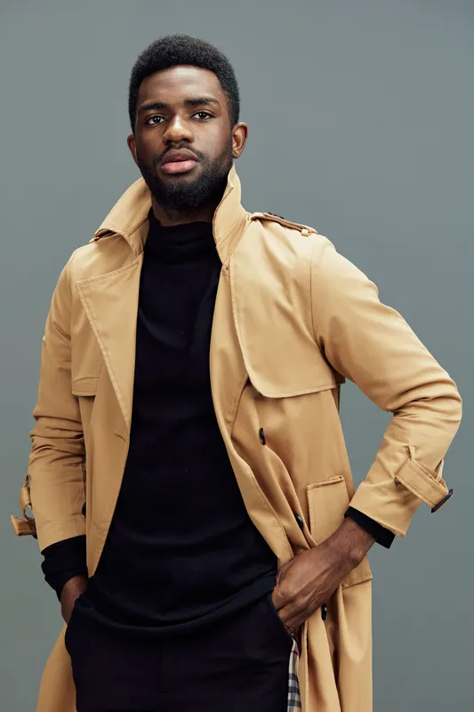 Young African man in a stylish beige trench coat, exuding confidence and sophistication against a muted gray background, perfect for fashion and lifestyle themes
