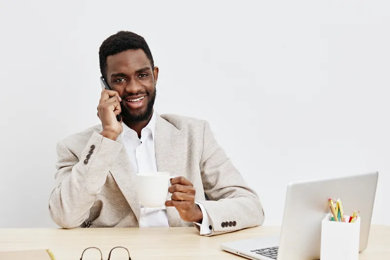 Happy smiling man with dark skin talking on mobile phone while sitting at desk with cup of coffee and laptop isolated on plain white background. Business, communication, technology concept.