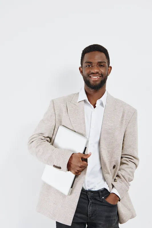 Smiling confident young African American man dressed in beige blazer and white shirt holding laptop, standing against plain white background, studio portrait, professional, business, fashion, confiden