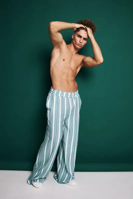 Young man posing shirtless with hands on head wearing striped wide pants against green background in studio. Fashion, casual style and confident expression.