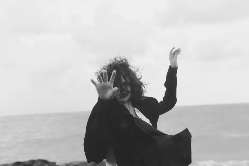 Dynamic movement in a wind swept scene, a flowing coat and raised arms against a seascape, conveying energy, freedom, and motion on a gray beach.