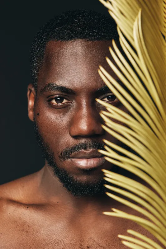 African man with dark skin and beard posing behind yellow palm leaf in creative conceptual visual style. Closeup portrait with serious expression and artistic lighting on black background.