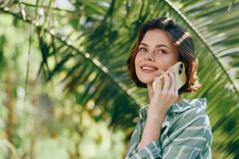 Smiling woman outdoors chats on a phone amid lush tropical greenery and warm sunlight.