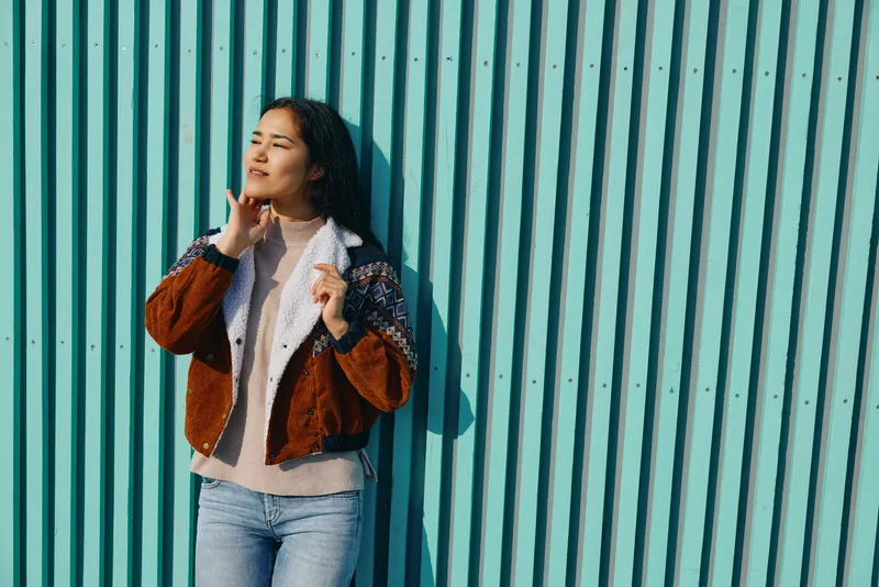 Woman leaning against a turquoise striped wall, wearing a brown patterned jacket and denim jeans, posing with a thoughtful gesture in daylight outdoors