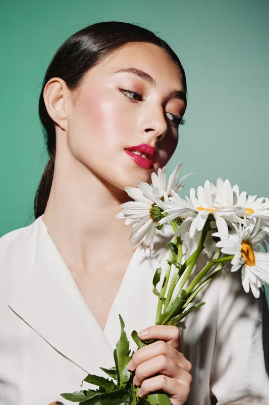 Young woman with sleek hair and vibrant pink lips holding white daisies wearing a white blazer against a soft green background. Concept of beauty, fashion, and floral creativity.