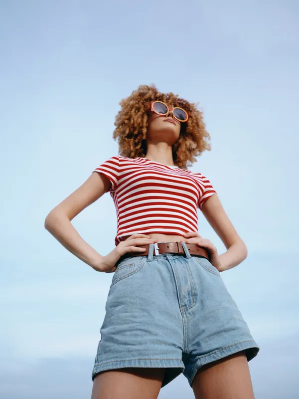 Person stands tall in a sunny scene wearing a striped red and white t shirt, denim shorts, and a belt, exuding confidence and carefree fashion against a bright blue sky.