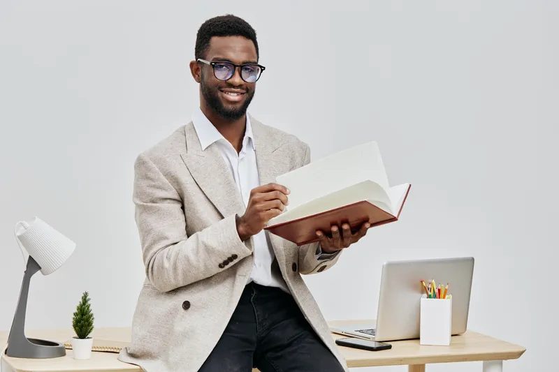 Smiling man with glasses and dark curly hair reading an open book in a modern office setting, casual business attire, relaxed mood, minimalistic decor, natural lighting, professional environment, indo