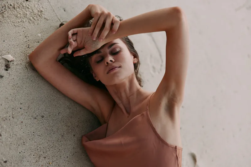 Serene portrait of a woman resting against a textured wall, arms raised, eyes closed, wearing a soft peach top, conveying calm, vulnerability, and quiet strength.