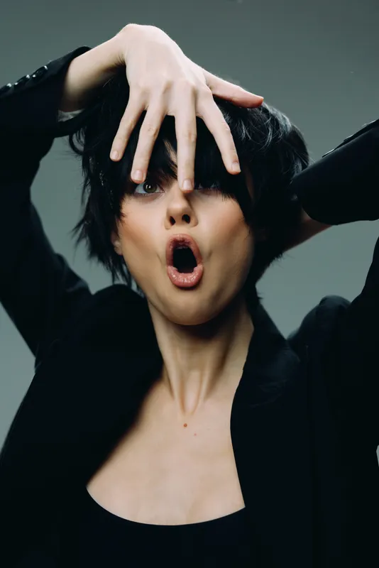 expressive woman in black jacket making dramatic gesture with hand over face, artistic portrait, dark background