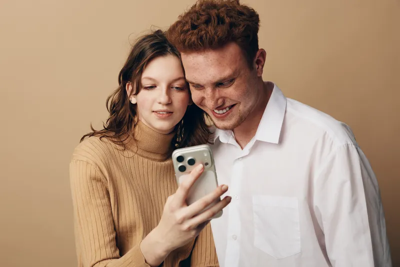 Young couple with casual clothes smiling and looking at smartphone screen together. Indoor portrait on beige background. Concept of friendship, technology, and happy moments.