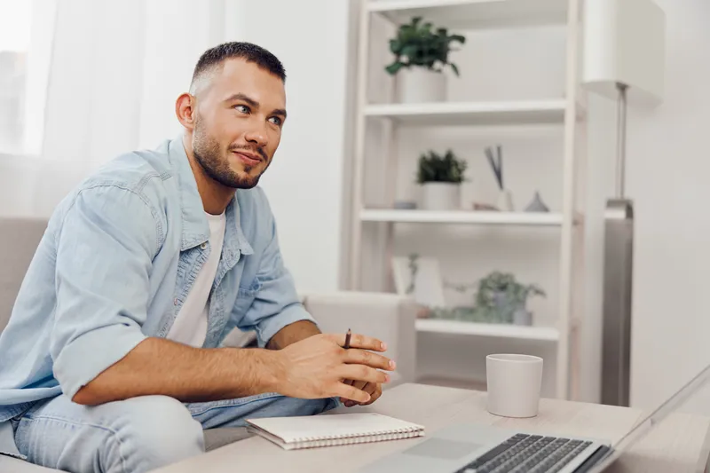 Thoughtful man sitting on a cozy couch, holding a pen and notepad, contemplating ideas in a bright and modern living room filled with green plants.