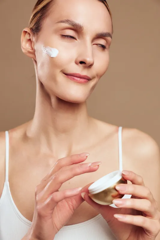 Young woman with clear skin applying white face cream on cheek with closed eyes. Concept of skincare, beauty routine, wellness, natural care, and self-care. Soft lighting.