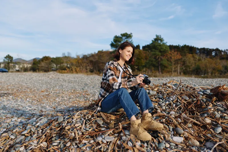 Woman sitting on a pile of leaves by a shoreline, wearing a plaid shirt and jeans, holding a camera and smiling while enjoying outdoor photography in autumn light