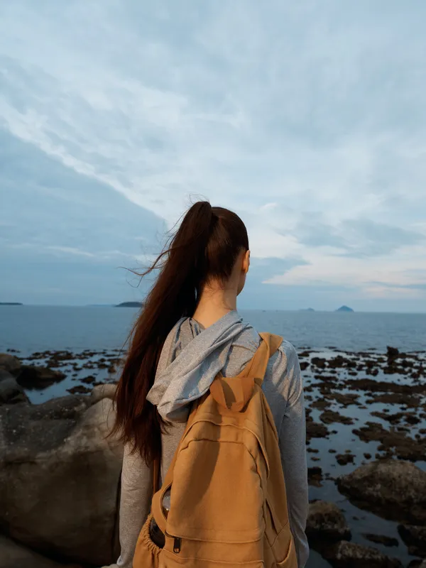 Woman with long hair in a ponytail gazing at the ocean, wearing a casual hoodie and backpack, evoking a sense of adventure and contemplation near the shore
