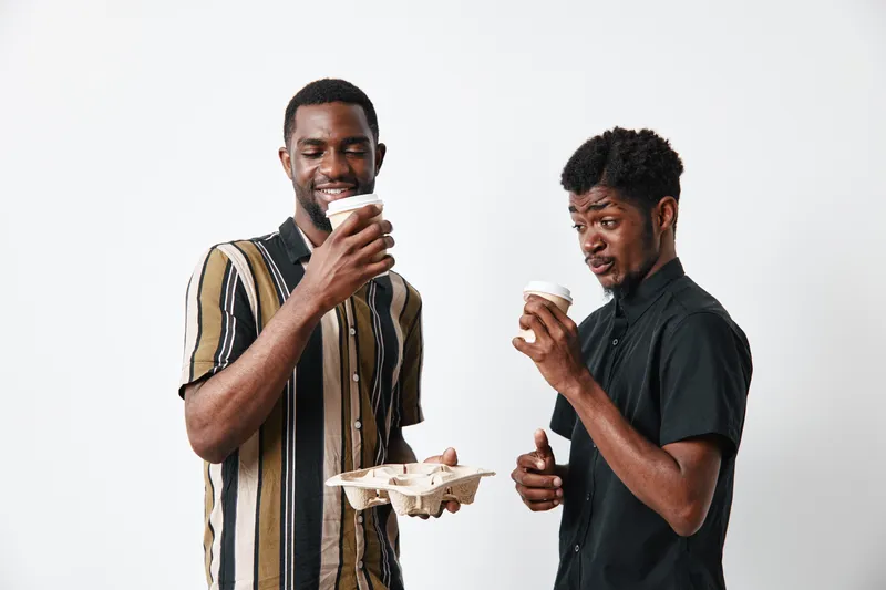 Two african men with dark skin holding coffee cups and cardboard tray standing on white background. Casual clothing, friendship and relaxed mood with beverage and smile.