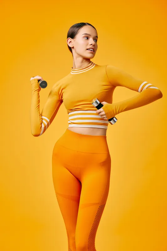 Fitness woman in a bright orange sport set holding a dumbbell, mid-action pose in a vibrant studio. Sporty posture, focused energy and dynamic training scene emphasizing health and performance.