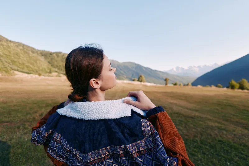 Woman in patterned jacket standing in meadow outdoors, back portrait gazing at mountain range, autumn light, serene landscape, nature travel and contemplative moment.