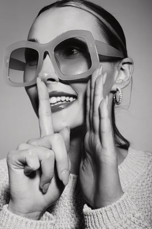 Happy Caucasian woman with light skin wearing large sunglasses and sweater gestures silence with finger near lips. Studio portrait in black and white.