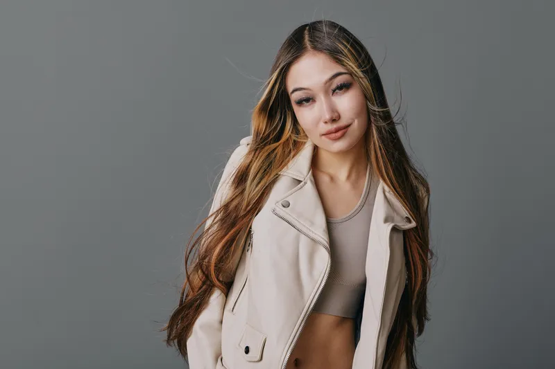 Young woman with long hair posing against gray background wearing light jacket and crop top, looking confident and stylish with natural makeup in studio portrait.