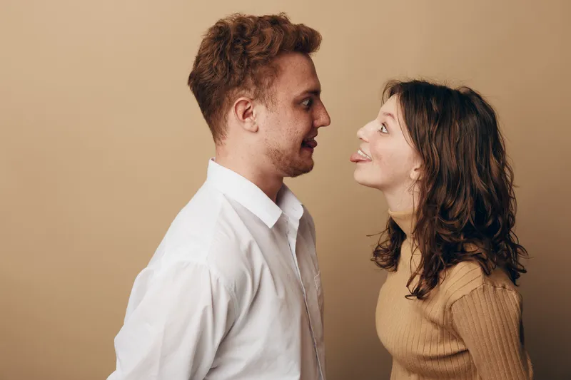 Young man and woman face each other playfully, woman sticking out tongue. Casual clothing and neutral background create a lighthearted, fun, and spontaneous moment between couple.