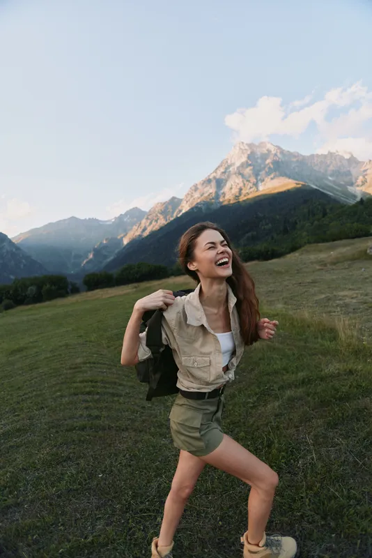 Woman hiking in the mountains, outdoor lifestyle scene, wearing beige jacket and shorts, carrying backpack, smiling at expansive alpine scenery, sunny day, natural landscape, candid travel moment