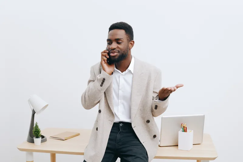 Confident young man with dark skin talking on mobile phone in modern office setting, wearing smart casual outfit, holding hand in gesture, professional workspace background with desk and laptop, lifes