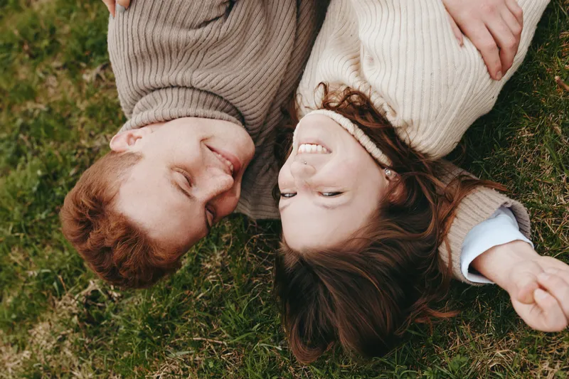 Young couple in cozy sweaters lies on grass smiling joyfully enjoying a relaxed autumn moment in nature with warm casual clothing and soft natural light
