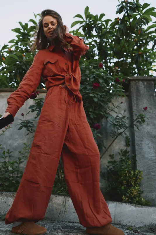 A woman in a rust red jumpsuit walks outdoors, posing with confident movement. Bright daylight highlights fabric texture in a casual lifestyle moment
