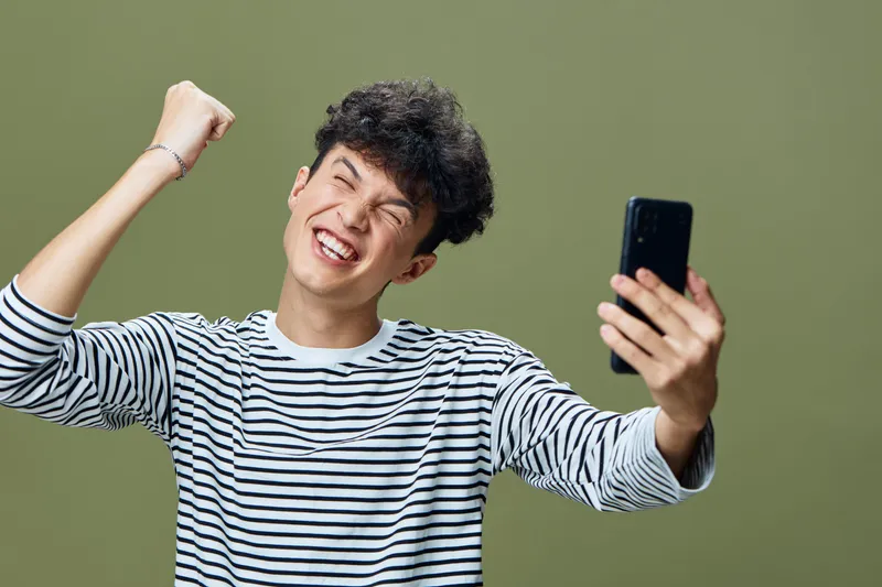 excited young man taking selfie with smartphone, wearing striped shirt, smiling broadly, raised fist, olive green background, casual moment of joy and success