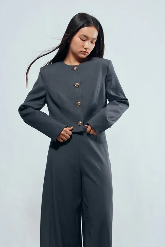 Woman in dark gray formal suit with long jacket and pants standing against light background. Young female model with straight hair styled naturally, looking down with calm expression.