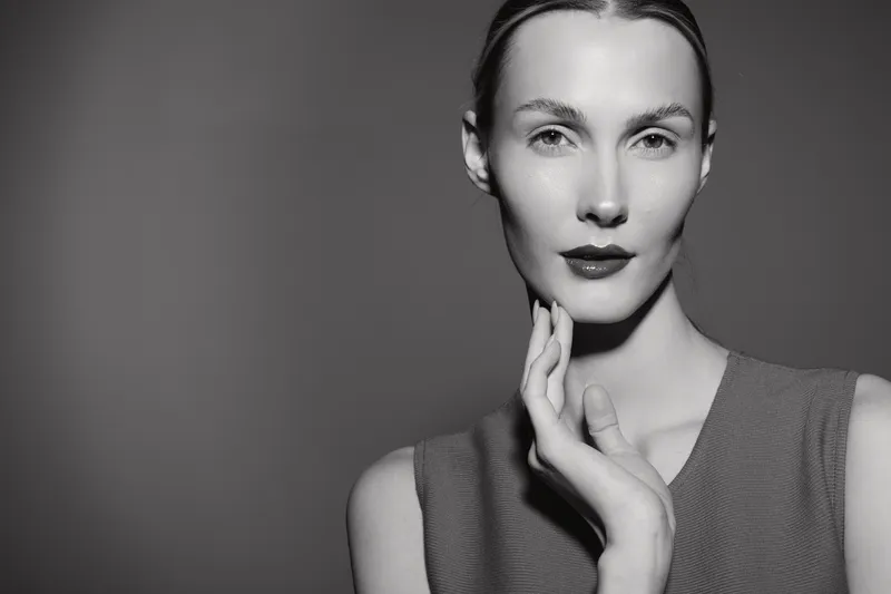 Portrait of a fair skinned Caucasian woman with slicked back hair touching her chin in a studio. The monochrome image highlights elegance and beauty in a minimalistic setting.
