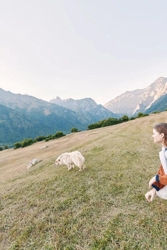 Dog and woman on grassy meadow near mountains during a hiking trip, enjoying nature and wide landscape as the pet explores the grass while the person watches in a calm outdoor scene.