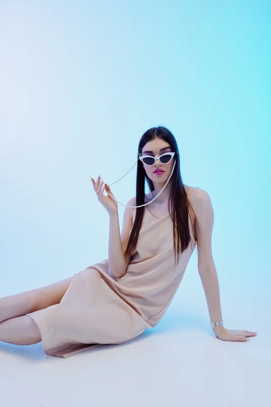 Stylish woman beauty fashion model in beige dress and white sunglasses poses sitting on floor against blue background. Concept for elegance, style, and modern trends.