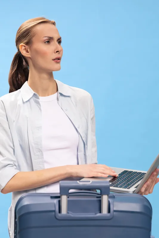 Young woman in white shirt holds laptop and blue suitcase, looking sideways on blue background. Concept for travel, business trip, and remote work with luggage and technology.