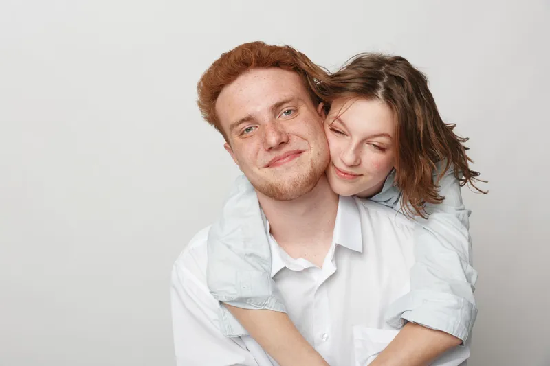 Smiling young couple with woman hugging man from behind on white background. Happy affectionate relationship, love, friendship, and casual lifestyle concept with natural expressions.