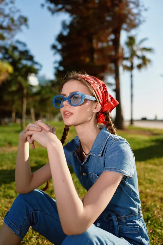 Young woman wearing blue sunglasses and a red headscarf with braids sitting outdoors in casual denim outfit on sunny day in park setting.