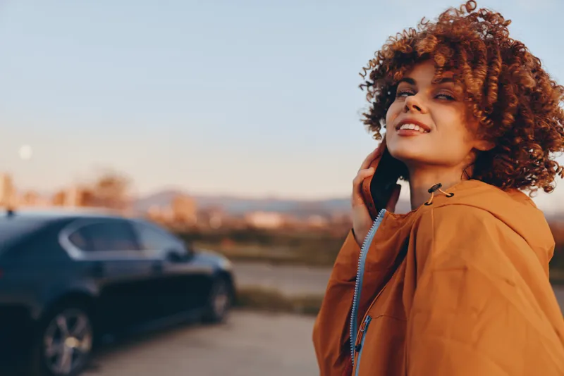 Woman with curly hair holding a phone by a car, outdoor street call with a smile. Lifestyle during sunset traffic, warm light, soft bokeh background, calm city road air calmness.