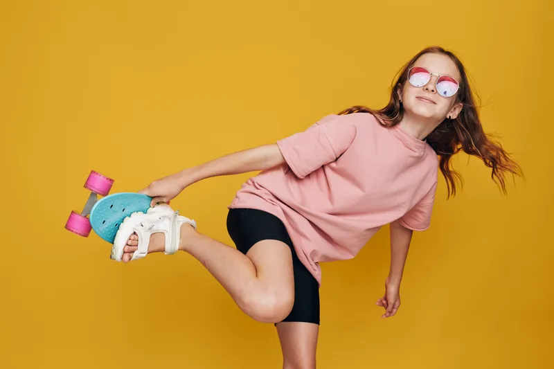 Smiling girl in pink t-shirt and black shorts holds a blue roller skate, posing against a yellow background. She wears round sunglasses and has long hair.