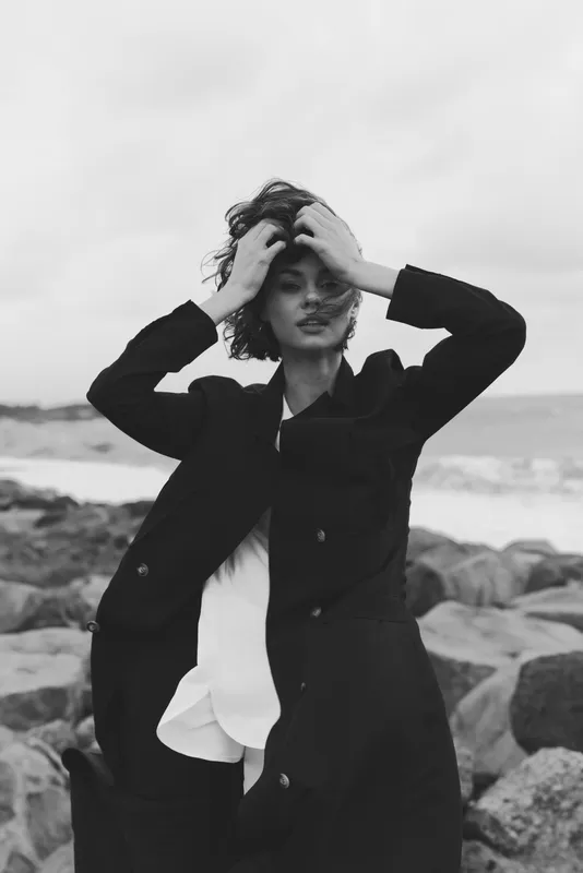 Fashion and mood on the coast with a stylish coat, wind in the hair, hands on the head. Black and white photography captures seaside drama and a confident pose.