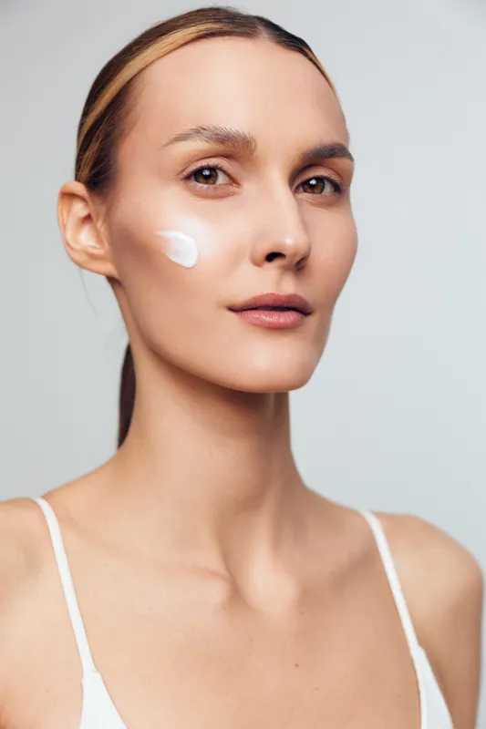 A young woman with clear skin applies white cream on her cheek, looking confidently at the camera against a plain light background. Concept for skincare, beauty, and self-care.