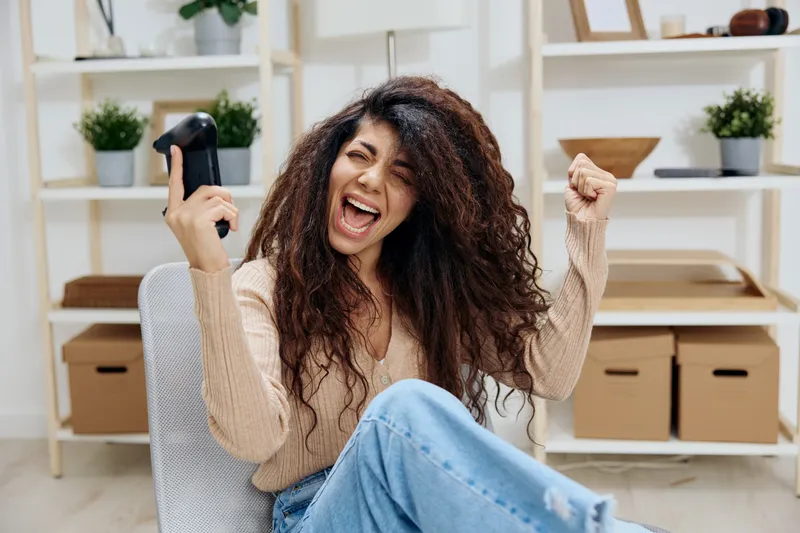Невероятно счастливая, везучая латиноамериканка с загаром и вьющимися волосами, победительница, поднимает руки вверх с джойстиком и геймпадом, сидит на стуле в современном домашнем интерьере, закрыв…