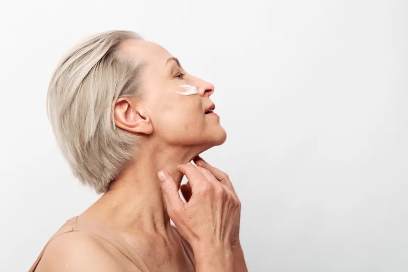 Mature woman with short grey hair applies white face cream on cheek and neck, eyes closed in calm studio. Concept of skincare, anti aging, self care, and natural beauty for older skin.
