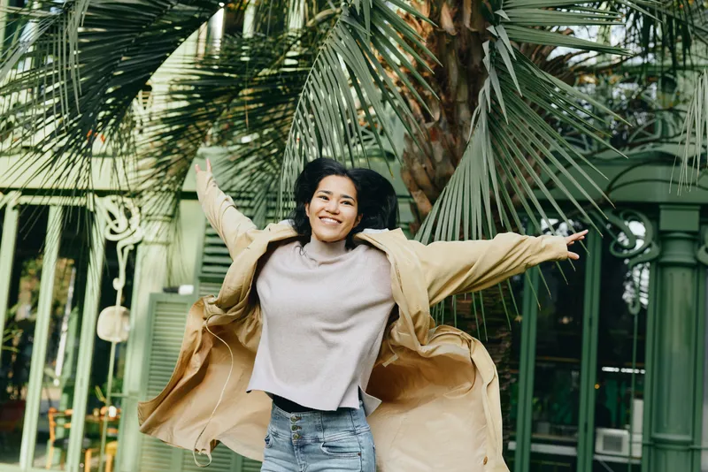 Joyful outdoor moment with a smiling woman arms outstretched beside lush palm leaves, wearing a beige coat and light sweater, capturing carefree energy and urban garden vibes and sunshine.