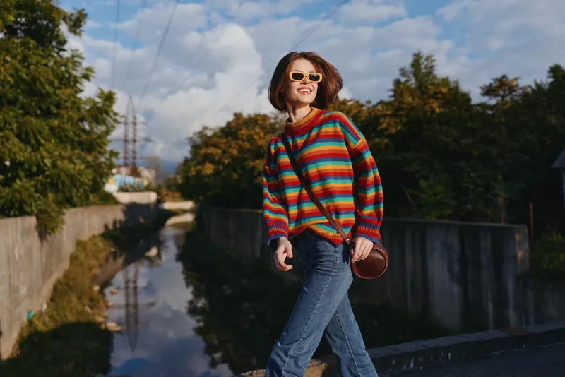 Woman in a colorful striped sweater wearing sunglasses and smiling while walking by a canal in urban nature, casual outfit with crossbody bag, joyful travel lifestyle scene.