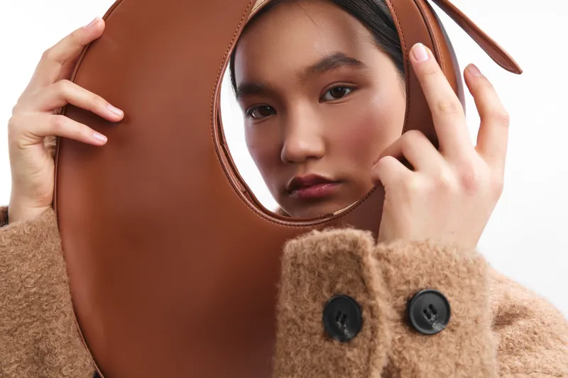 asian woman peers through a circular brown leather accessory, wearing a cozy textured coat. The close-up captures her curious, composed expression, delivering a stylish, artful mood.
